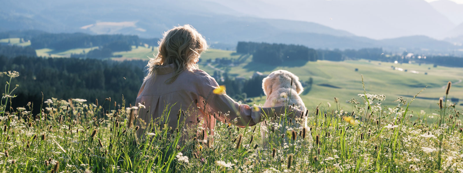 Katharina and Nala sit on a meadow and look down from a hill onto the meadow and woods below.