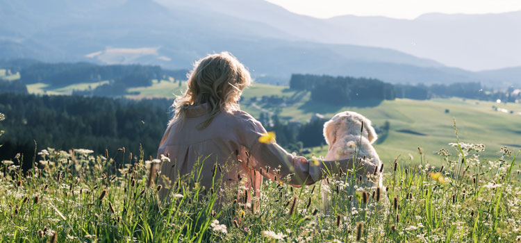 Katharina and Nala sit on a meadow and look down from a hill onto the meadow and woods below.